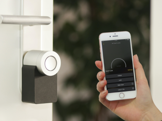 A hand holding a smartphone with a smart lock control app on the screen, next to a modern smart lock installed on a door.