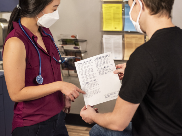 A healthcare professional in a maroon scrub and a masked individual in a black shirt reviewing a document together in a clinical setting.