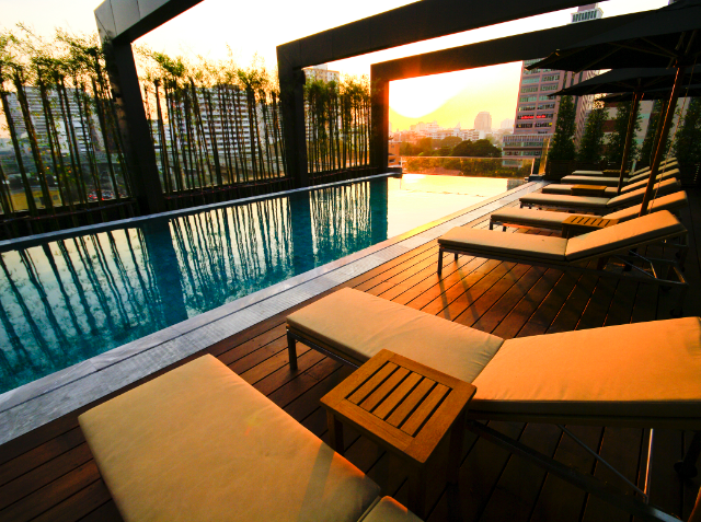 Outdoor swimming pool on a wooden deck with lounge chairs at sunset, overlooking a city skyline.