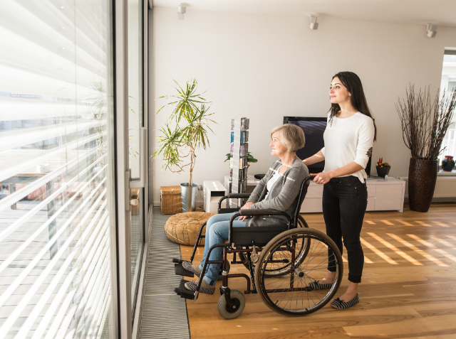 A younger woman standing behind an older woman in a wheelchair, both looking out of a large window in a modern apartment.