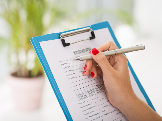 Close-up of a hand with red nail polish filling out a medical form on a clipboard with a pen, with a blurred indoor background.