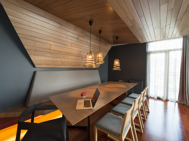 A modern dining or work space with a slanted wooden ceiling and wall, a long table with chairs and a bench, stylish pendant lights, and a laptop on the table. A large window with sheer curtains is on the right, and the opposite wall is dark gray.