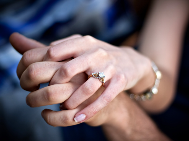 Two hands clasped together, one with a gold ring set with a large stone on the ring finger.