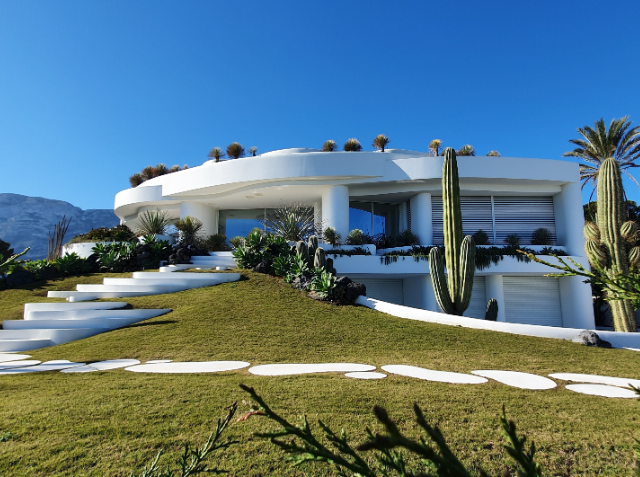 Modern white house with curved architecture and a green roof, surrounded by a landscaped garden with cacti and succulents, stone steps on a grass lawn, under a clear blue sky with mountains in the background.