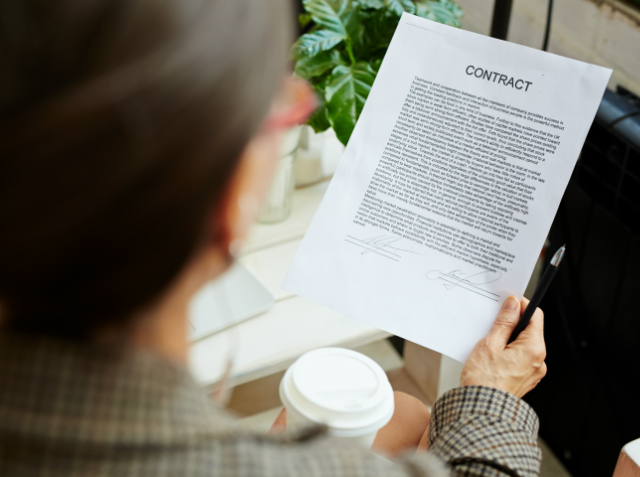 Over-the-shoulder view of a person reviewing a contract with signatures at the bottom, holding a coffee cup in the other hand.