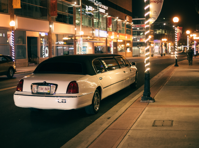 A white stretch limousine parked on a city street at night with illuminated storefronts and streetlights in the background.