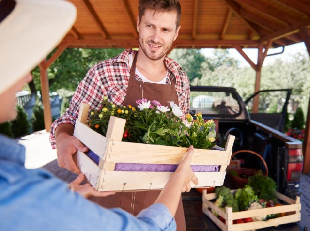 Smiling man in an apron handing over a crate of flowering plants to another person at an outdoor market.