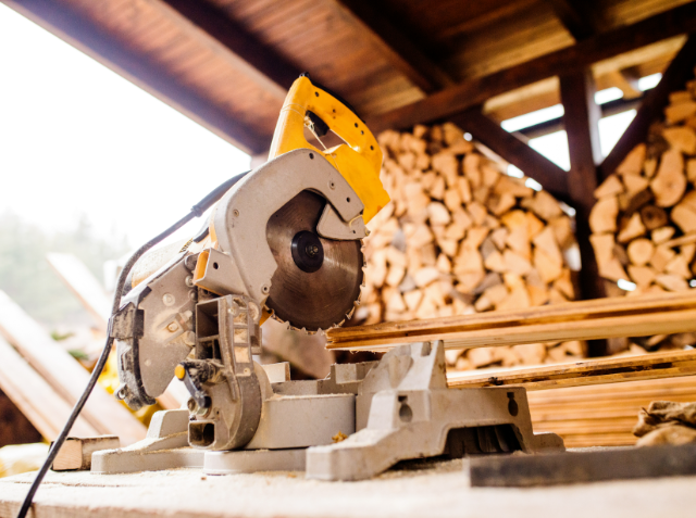 A miter saw on a workbench with wood pieces, in a workshop with a pile of firewood in the background.