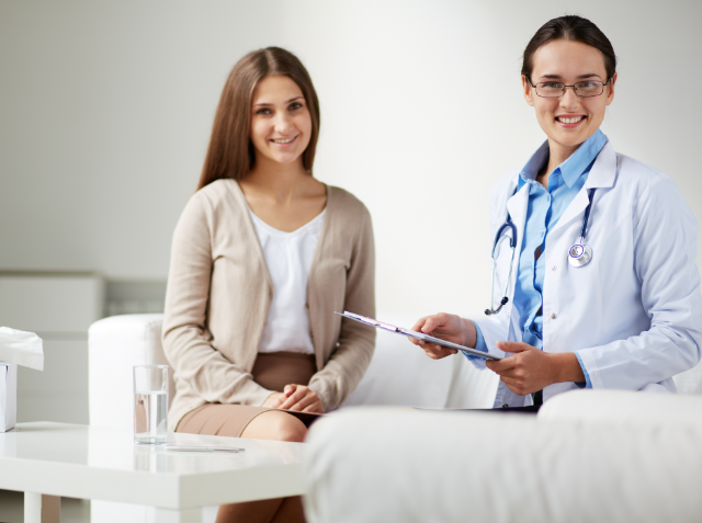 A smiling patient seated on a couch and a healthcare professional with a clipboard standing next to her in a medical office.