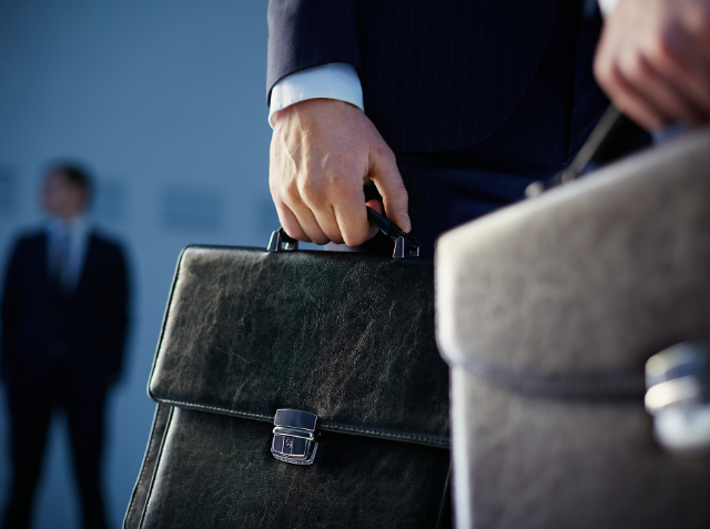 Close-up of a person's hand holding a leather briefcase, wearing a dark suit, with another individual in the background, indicating a business environment.