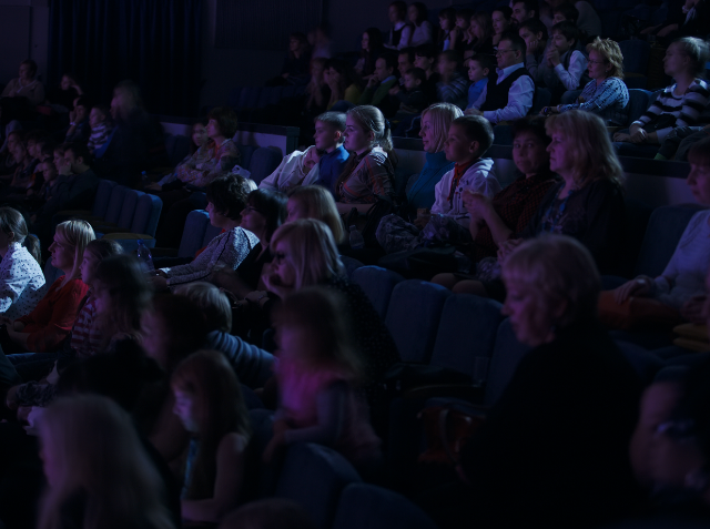 Audience of adults and children seated in a dark theater, attentively watching a performance or screen.