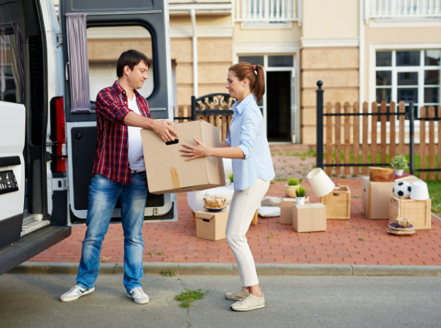 A man and a woman are transferring a cardboard box from a moving van to a house with more boxes and items on the ground, indicating a moving process.