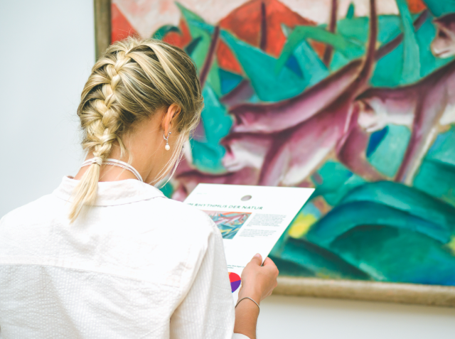 A woman with a braided hairstyle, seen from behind, is reading a brochure while observing an abstract painting in an art gallery.