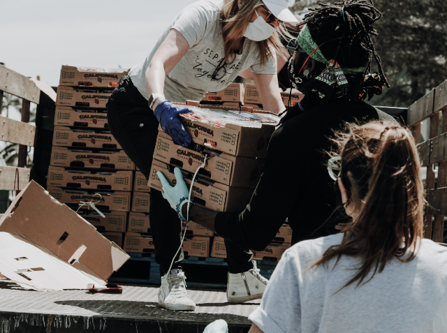 Two people wearing gloves and face masks are handling cardboard boxes during a volunteer supply distribution event, with stacks of boxes in the background.