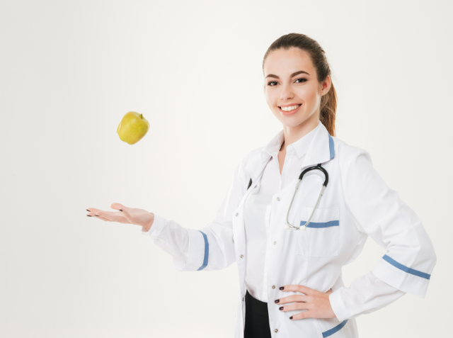 A smiling woman in a white lab coat with a stethoscope around her neck is tossing a green apple in the air.