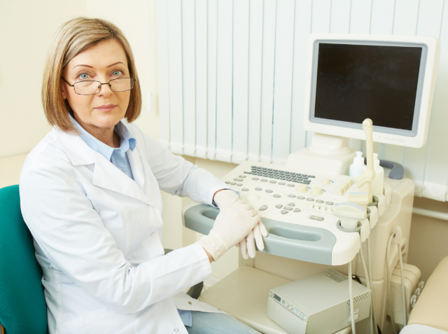 A female medical professional in glasses and a white lab coat sitting at an ultrasound machine.