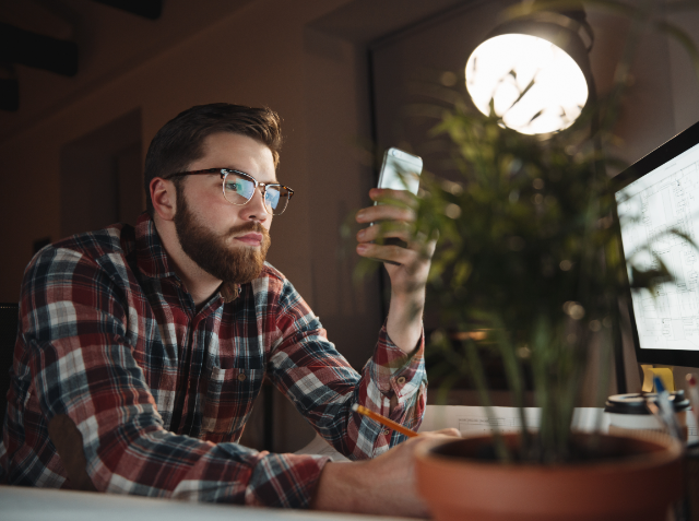 Bearded man in glasses examining his smartphone with focus, sitting at a desk with a computer monitor and a potted plant, in a dimly lit room.