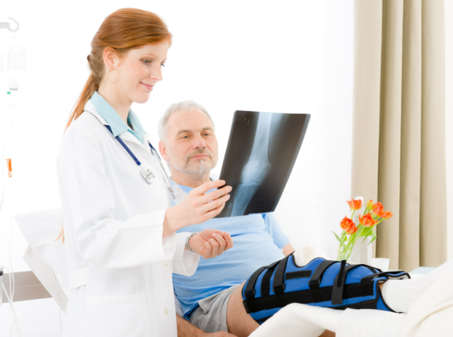 A medical professional examining an X-ray film next to a seated older man with a knee brace in a medical office.