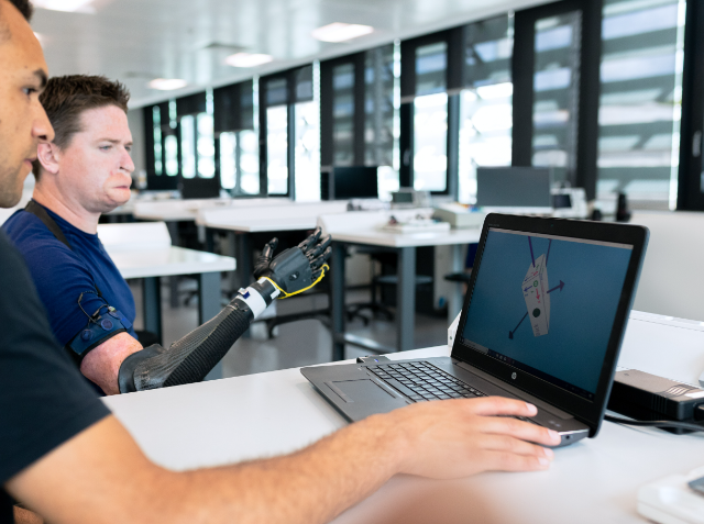 Two men at a desk with a laptop displaying a 3D model of a prosthetic limb; one man has a bionic prosthetic arm and is collaborating with the other man.