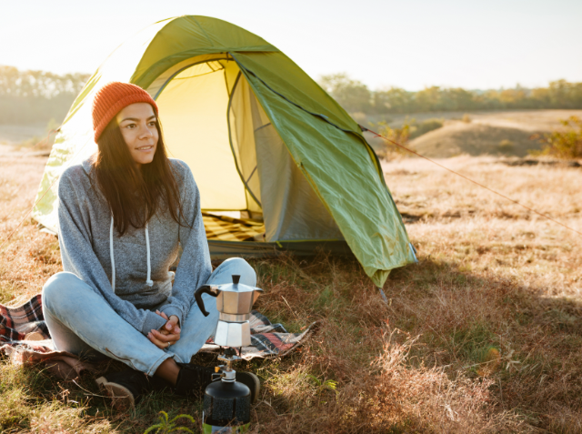 A woman in a red beanie and grey sweatshirt sits on a blanket outside a tent with a moka pot on a camping stove, in a grassy field during a sunny day.