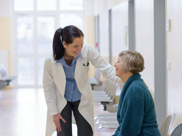 A healthcare professional in a white coat smiling at an older seated woman in a teal sweater in a bright clinical hallway.