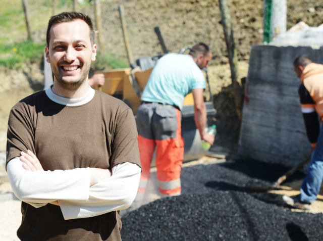 A smiling man with crossed arms in the foreground, with two workers laying asphalt in the background on a sunny day.