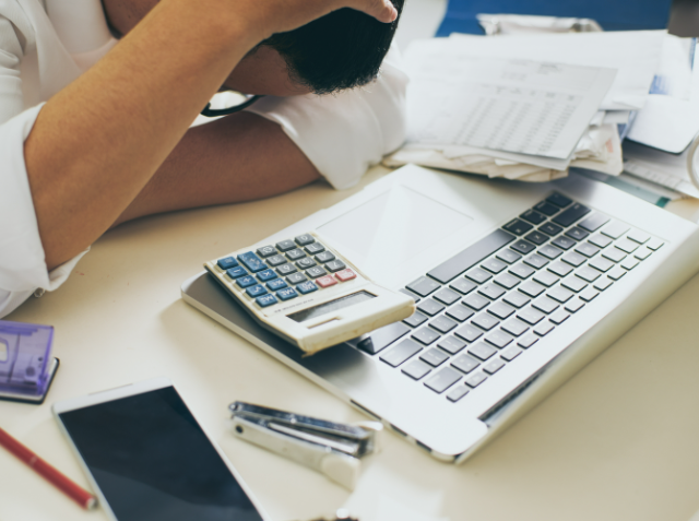 Stressed individual at a desk with a laptop, calculator with a credit card on it, mobile phone, and scattered papers, indicating financial stress or work overload.