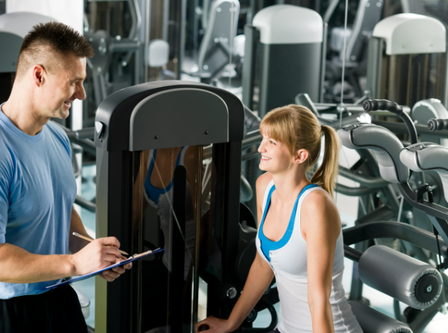 A personal trainer with a clipboard discussing exercise with a female client in a gym surrounded by fitness equipment.