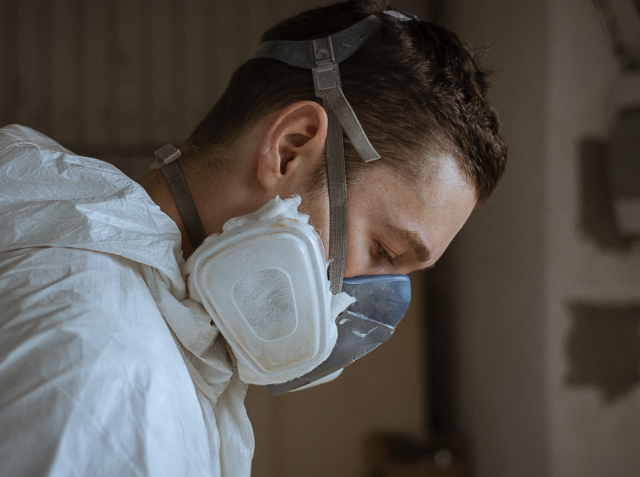 Person in a white protective suit and respirator mask with eyes closed, in a moment of rest or contemplation.