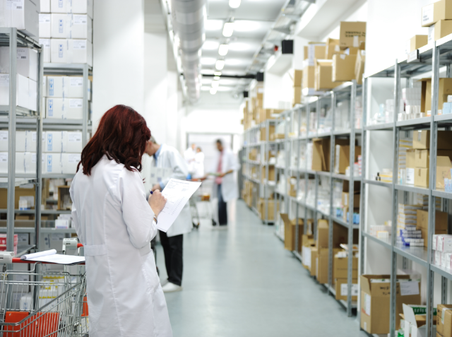 A person in a white lab coat reviews paperwork in a medical or pharmaceutical supply warehouse with shelves stocked with boxes.