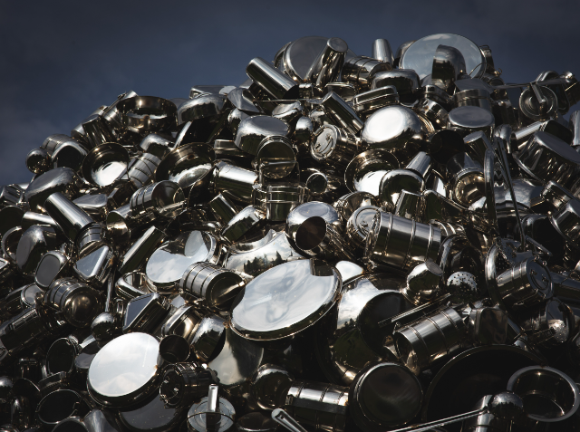 A large pile of shiny stainless steel kitchen pots and pans with various sizes and shapes, reflecting light under a cloudy sky.