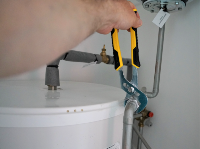 A person's hand using an adjustable wrench on a pipe fitting connected to a water heater, with various plumbing fixtures in the background.