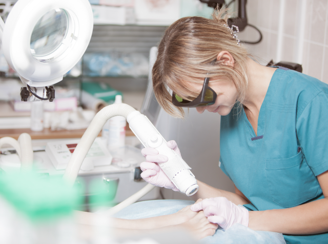 A healthcare professional in teal scrubs and protective glasses using a handheld laser device on a patient's skin in a clinical setting.