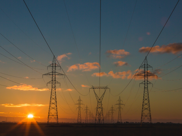 Silhouetted electricity pylons in a row at sunset with power lines and a few clouds in the sky.