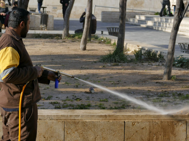 A man in a brown jacket and yellow vest using a hose to spray water on a paved surface in a park setting.