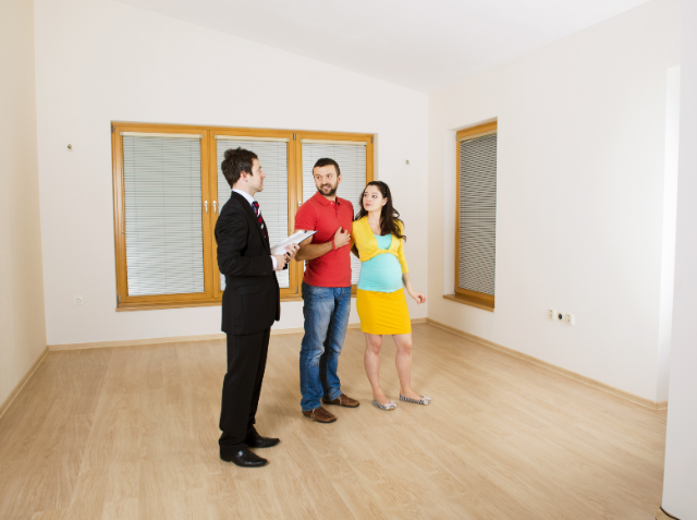 A real estate agent in a suit holding papers discusses with a couple, with the woman being pregnant, inside an empty room with hardwood floors and large windows.