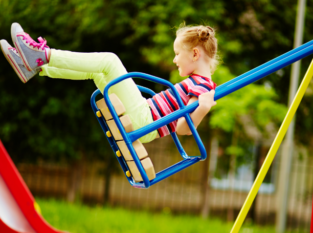 A young girl with her hair in a bun, wearing a striped shirt and green pants, joyfully swings on a blue and yellow swing set at a playground with green grass and a red slide in the background.