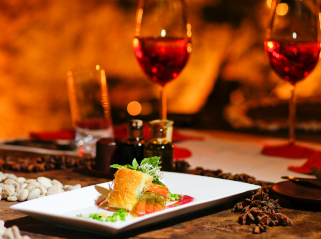 A romantic dinner setting with a gourmet dish in the foreground, two glasses of red wine, and a glowing fireplace in the background, creating a cozy atmosphere.