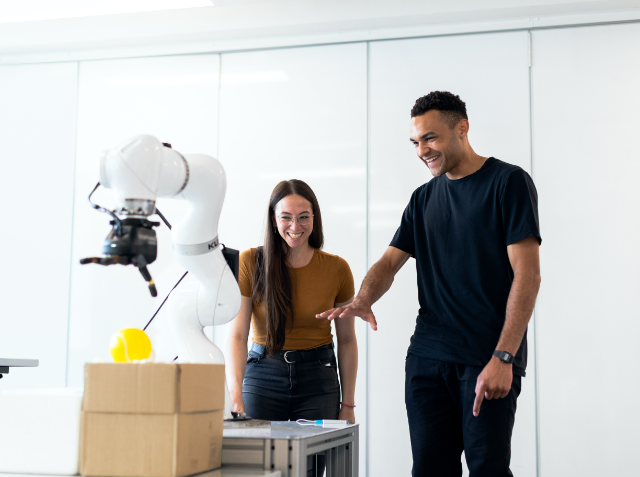 A man and a woman interacting with a robotic arm in a laboratory, with the robot holding a yellow object, both individuals appear fascinated and pleased.