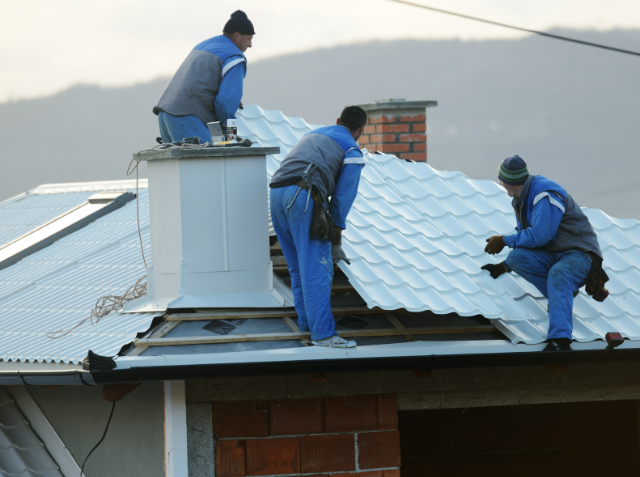 Three workers in blue uniforms installing or repairing a corrugated metal roof on a building, with hills in the background.