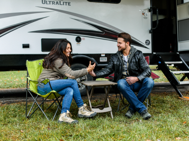 A man and a woman sitting on folding chairs by an 'ULTRALITE' travel trailer, smiling and conversing in a camping setting.