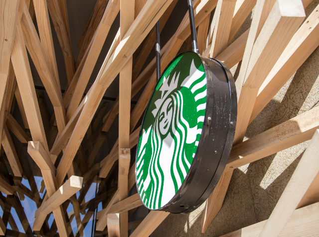 Starbucks logo on a round, illuminated sign mounted on a wall with an overhanging wooden beam structure.