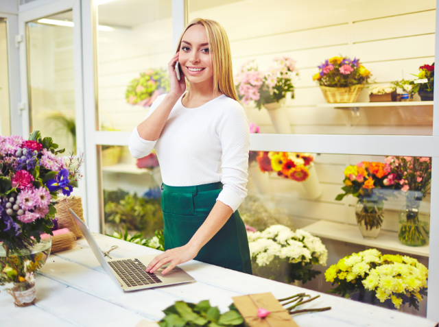 A smiling woman in a white top and green apron talking on a phone and using a laptop at a flower shop counter, surrounded by colorful flowers.