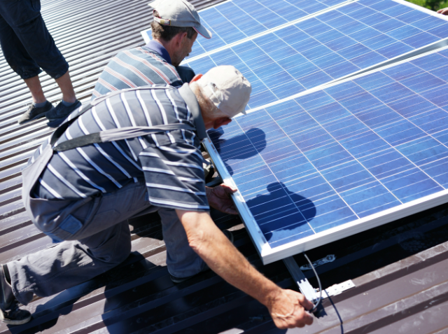 Two workers installing solar panels on a corrugated metal roof under a clear blue sky.