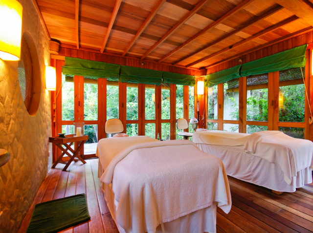 A tranquil massage room with two massage tables, wooden beams, stone walls, and large windows overlooking greenery.