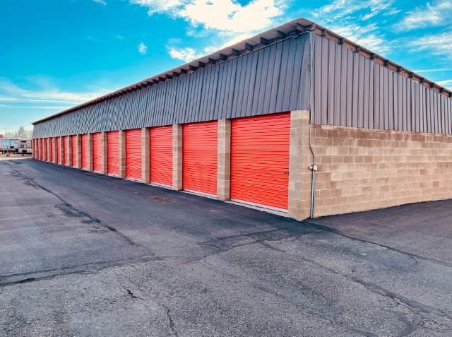 A single-story storage facility with a row of red roll-up doors under a gray metal roof, against a blue sky with sparse clouds.