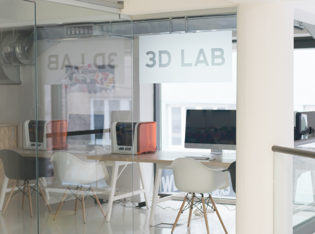 A 3D printing lab with two 3D printers on a table, white chairs, and a computer monitor, viewed through a glass wall with '3D LAB' written on it.