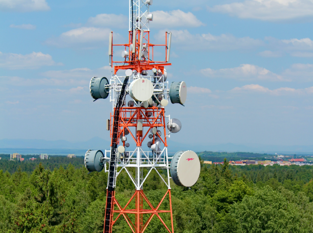 A tall telecommunications tower with white and orange stripes, equipped with antennas and dishes, stands against a backdrop of a green forest and a clear blue sky, with a distant town on the horizon.