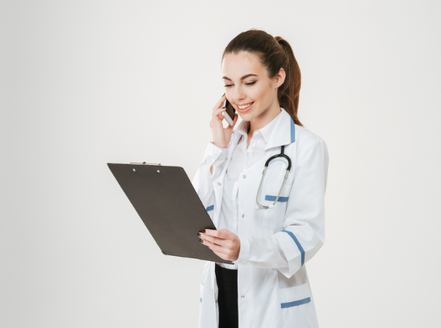 Smiling female medical professional in a white lab coat with a stethoscope, looking at a clipboard while talking on a mobile phone.