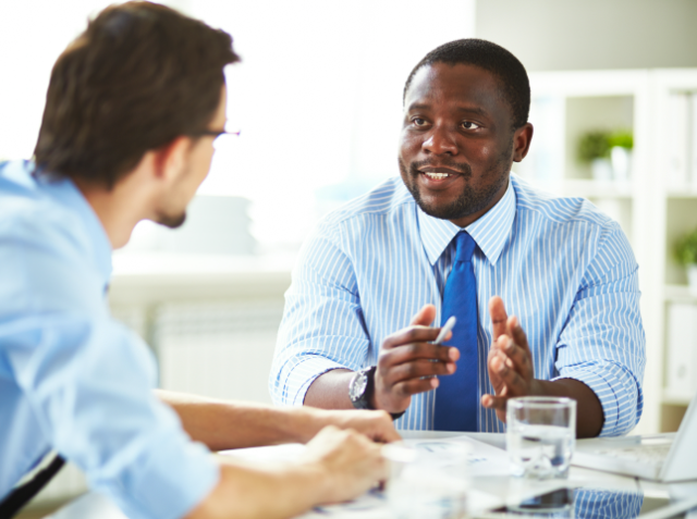 Two men in a business meeting, one gesturing while talking and the other listening, with a laptop and papers on the table.